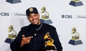 Khris Riddick-Tynes smiles, holding a Grammy award and pointing to it. He wears a black cap and jacket.