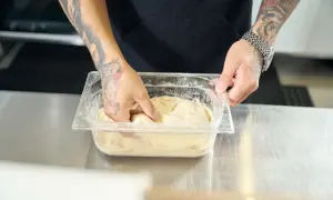Ręce z tatuażami mieszają ciasto na pizzę w pojemniku. Poznaj prosty sposób, jak rozmrozić ciasto na pizzę, by było idealne.