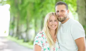 Ett leende par, Nanna Olasdotter Hallberg och hennes partner, njuter av en sommardag utomhus bland gröna träd.
