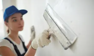 Kobieta w niebieskiej czapce i rękawiczkach szlifuje białą ścianę gładzią.