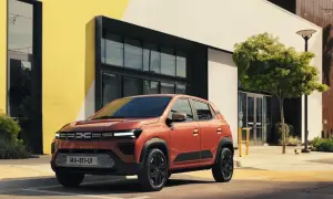Pomarańczowy Dacia Spring Electric zaparkowany przed nowoczesnym budynkiem z żółtymi akcentami.