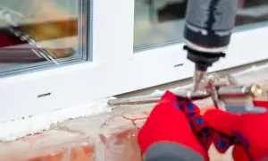 Uszczelnianie okna pianką montażową. Ręce w czerwonych rękawiczkach trzymają pistolet, aplikując piankę na zimny parapet wewnętrzny.