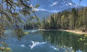 Spiegelglatter See mit Bergpanorama in den bayerischen Alpen. Nadelbäume säumen das Ufer, im Hintergrund ragen schneebedeckte Gipfel auf.