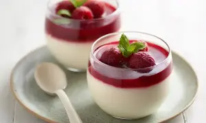 Två glas med krämig pannacotta med hallonspegel, toppad med färska hallon och mynta. En ljus sked ligger på fatet.