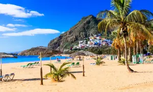 Plage de sable doré, palmiers, parasols et maisons colorées perchées sur une colline. Une invitation à visiter Tenerife pour des vacances de rêve.