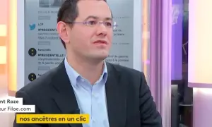 Un homme en costume, lunettes, parle. Le nom de famille gratuit "Filae.com" est visible, suggérant une recherche d'ancêtres.