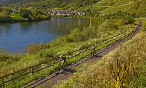 Zwei Radfahrer genießen die idyllischen radwanderwege deutschland entlang eines Flusses mit Weinbergen und einem Dorf im Hintergrund.