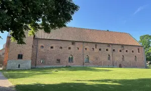 Imponerande tegelbyggnad med tegeltak, en del av Vadstena kloster, omgiven av grönskande gräsmatta och träd under en klarblå himmel.