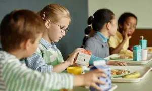 Kinder essen in der Mensa. Ein Mädchen mit Brille hält eine Milchpackung. Ein Teil des Tagesablaufs eines Schulbegleiters.
