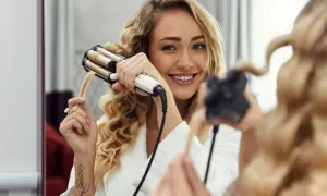 Uśmiechnięta blondynka w szlafroku tworzy piękne fale na włosach za pomocą karbownicy.