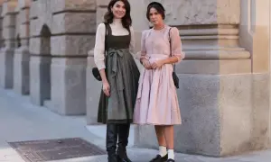 Zwei Frauen posieren vor einer Steinfassade. Eine trägt ein grünes Dirndl mit weißer Bluse und schwarze Stiefel.
