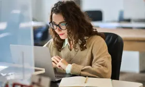 Donna con occhiali lavora al laptop, concentrata. Forse sta cercando la miglior surroga per un progetto.