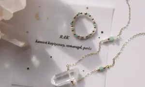 Złota biżuteria z kryształem, perłami i zielonymi kamieniami. Napis "Rak" i "kamień księżycowy, szmaragd, perła" sugerują, że to kamień zodiakalny dla Raka.