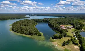 Malownicze jezioro Woświn otoczone lasem, z hotelem i polem kempingowym na brzegu. Błękitne niebo z białymi chmurami.