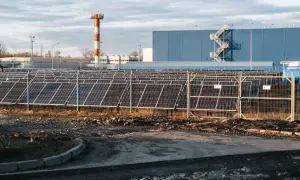 Duże pole paneli słonecznych przed niebieskim budynkiem. W tle pomarańczowo-biała wieża. THD.