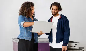 A woman and man review a laptop, discussing music artist management. They stand near stage equipment.