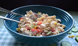 En stor blå skål fylld med krämig pastasallad med kyckling, tomat och majs. Två gafflar ligger ovanpå.