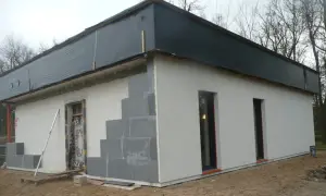 Budowa domu ze styropianu w toku. Ściany z bloczków, częściowo otynkowane, dach przykryty czarną folią.