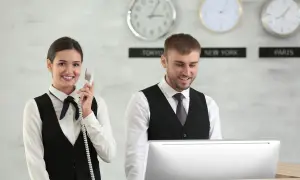 Uśmiechnięta recepcjonistka rozmawia przez telefon, podczas gdy kolega pracuje przy komputerze. Zegar pokazuje, ile godzin pracuje recepcjonistka w hotelu.