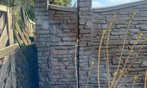Betonowy płot z fakturą kamienia, z widocznymi słupkami i fragmentem starego płotu.