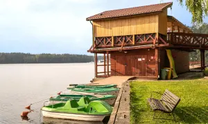 Zielone łódki przy brzegu, drewniana przystań i ławka na trawie. W tle jezioro i las, przypominające wrzosową górę.