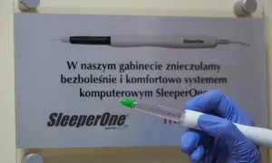 Rękawiczka w niebieskiej rękawiczce trzyma strzykawkę z zieloną końcówką, gotową do podania lidokainy. Tablica informuje o bezbolesnym znieczuleniu.