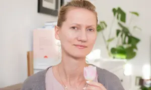 Kobieta bez makijażu, z delikatnym uśmiechem, trzyma w dłoni różowego tulipana. W tle widać rośliny i książki.