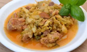 Pyszny bigos z młodej kapusty z kawałkami mięsa, podany na białym talerzu, ozdobiony gałązką bazylii.