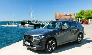 Mörkgrå Mazda CX-60 laddhybrid parkerad vid en brygga med båtar och en röd bil i bakgrunden. En solig dag för ett Mazda CX-60 laddhybrid test.