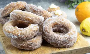 Un mucchio di ciambelle di carnevale zuccherate, alcune con crema, su un tagliere di legno, con arancia e limone sullo sfondo.