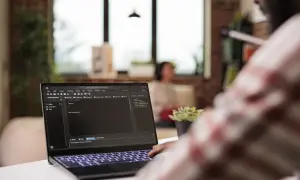 Programista pracuje nad kodem na laptopie, gdzie widać plik hosts. W tle siedzi osoba.