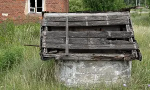 Zrujnowana, biesiadna kapela przy głębokiej studzience. Drewniany dach zniszczony, obok ceglany budynek.