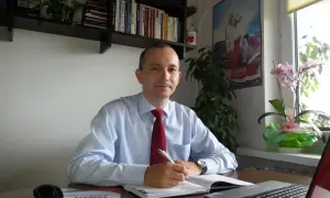 Mężczyzna w koszuli i krawacie siedzi przy biurku, pisząc w notatniku.