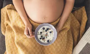 Ciąża i ochota na kwaśne. Kobieta w ciąży je jogurt z jagodami i granolą, otulona żółtym kocem.