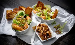 Zwei Schüsseln mit Hot Dog Salat, garniert mit Gurkenscheiben, geröstetem Brot und knusprigen Zwiebeln.