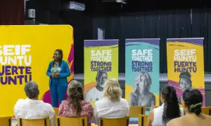 Een spreker staat voor een gele banner met "SEIF HUNTU FUERTE HUNTU". Achter haar staan banners met "SAFE TOGETHER STRONG TOGETHER". Dit is een evenement voor hss sw jeugd.