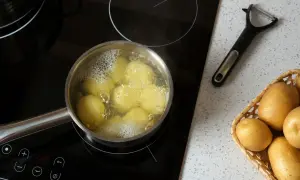 Ziemniaki gotują się w garnku na płycie indukcyjnej. Obok leży obieraczka i koszyk z surowymi ziemniakami. To początek, jak ugotować ziemniaki.
