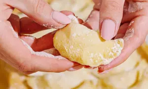 Dłonie z różowymi paznokciami formują ciasto do pierogów, obsypane mąką. W tle widać gotowe pierogi.