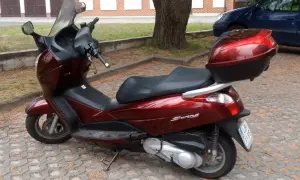 Ciemnoczerwony skuter Honda Swing 125 z czarnym siedzeniem i kufrem, zaparkowany na kostce brukowej.