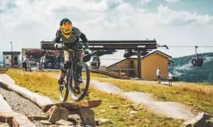 Rowerzysta pokonuje przeszkodę na drewnianej kładce w bike park Bielsko Biała. W tle kolejka linowa i budynek stacji.