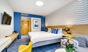 Pokój w hotelu Mistral Jurata: dwa łóżka, żółty fotel, kanapa, bukiet kwiatów i obraz z żaglówką.
