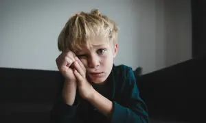 Chłopiec z blond włosami, zamyślony, opiera głowę na dłoniach. Jego spojrzenie sugeruje trudności, być może związane z zespołem Aspergera.