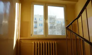 Słoneczne promienie padają na błyszczącą ścianę w klatce schodowej. Widać okno z widokiem na blok i kaloryfer.