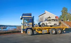 En blågul lastbil transporterar en vit Volvo Raske herrgårdsvagn längs en sjö.