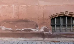 Wilgotne plamy na kamiennym murze piwnicy, wskazujące na potrzebę dowiedzenia się, jak osuszyć fundamenty. Widoczne kraty w małym oknie.