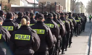 Policjanci w czarnych kurtkach z napisem "POLICJA" stoją w szeregu, gotowi do akcji. Widać, jak pracuje policja zmiany, przygotowując się do służby.