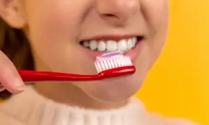 Dziecko z uśmiechem na ustach, zbliża czerwoną szczoteczkę z pastą do zębów. Czy fluoryzacja wybiela zęby?