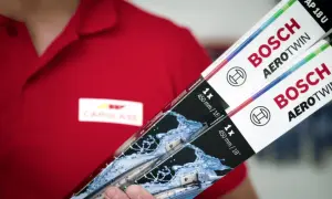 Person i röd pikétröja med Carglass-logga håller upp två paket med Bosch Aerotwin torkarblad.