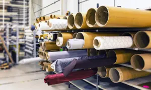 Różne rodzaje materiałów na rolkach, gotowe do użycia w produkcji. Od jasnych wzorzystych tkanin po ciemne, jednolite materiały.