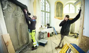 Två män arbetar med en stor stenrelief i en halvrund del av kyrka under renovering.